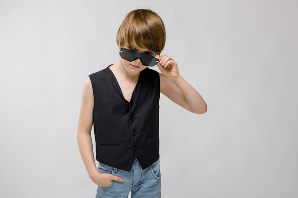 Cute caucasian boy in black waistcoat and sunglasses posing with different expressions on white wall in studio. - Foto, Bild
