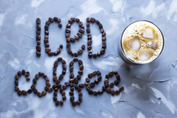 A glass of iced coffee with milk next to the inscription "ice coffee" from coffee beans on a gray concrete background. Summer cooling drink - Fotó, kép