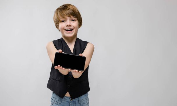 Cute chłopiec kaukaski w czarny strój, pokazując różne wyrażenia i trzymając smartfon na białej ścianie w studio. - Zdjęcie, obraz