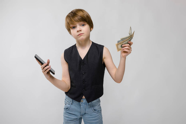 Cute chłopiec kaukaski w czarny strój, pokazując różne wyrażenia i trzymając smartfon z stosu dolarów na białej ścianie w studio. - Zdjęcie, obraz