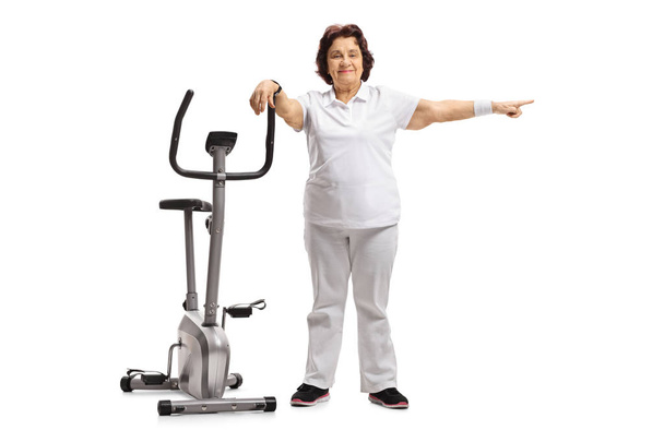 Full length portrait of an elderly woman leaning on an exercise bike and pointing isolated on white background - Foto, immagini