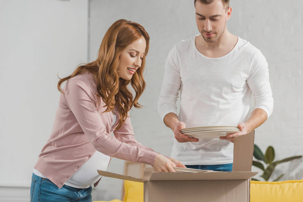 young pregnant couple packing plates in cardboard box during relocation - Photo, Image