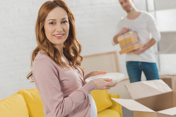 happy pregnant woman holding dishware and smiling at camera while relocating with husband - Photo, Image