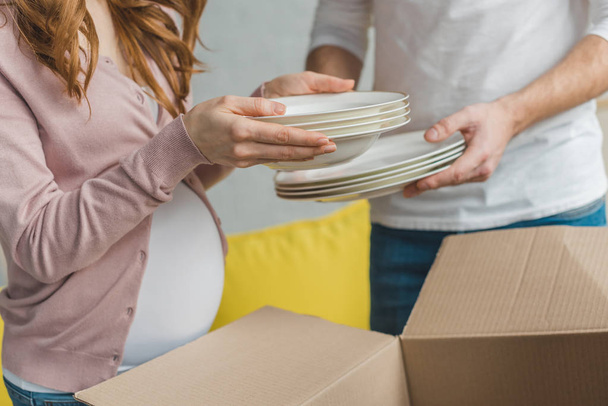 cropped shot of young pregnant couple packing plates in cardboard box during relocation - Photo, Image
