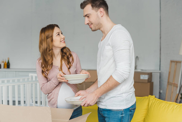 glückliches schwangere Paar hält Geschirr in der Hand und lächelt einander im neuen Haus zu - Foto, Bild