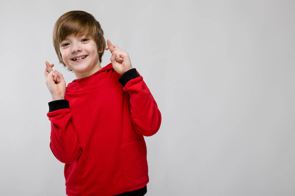 Nice preteen caucasian boy in casual outfit showing different expressions on white wall in studio. Smiling with crossed fingers. - Photo, Image