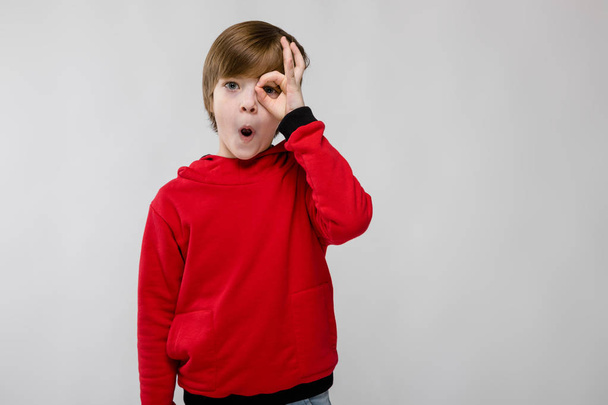 Nice preteen caucasian boy in casual outfit showing different expressions on white wall in studio. Looking through rounded fingers. - Photo, Image