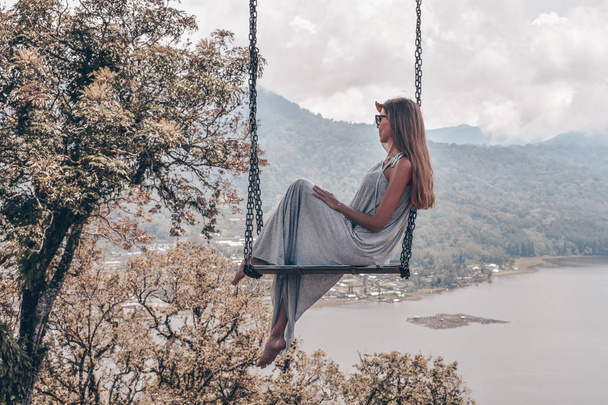 series traveling girl in Asia, beautiful girl with long dark hair in elegant grey dress sitting on the swing in beautiful nature place in Bali - Foto, Imagem