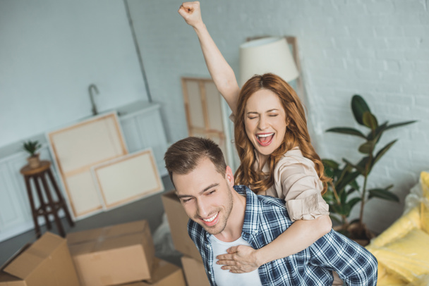 happy couple piggybacking at new apartment with cardboard boxes, relocation concept - Фото, изображение