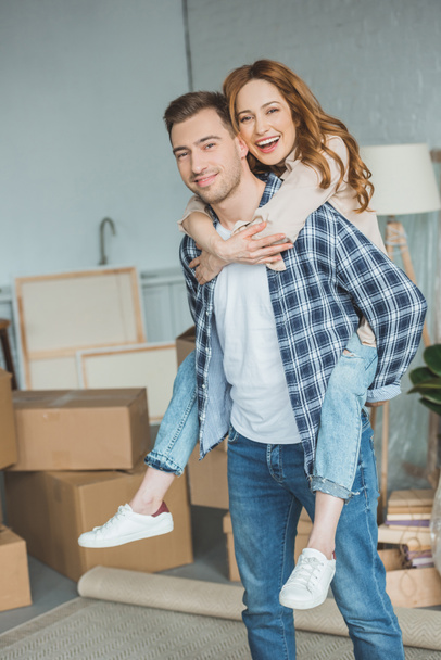 smiling couple piggybacking at new apartment with cardboard boxes, relocation concept - Fotoğraf, Görsel