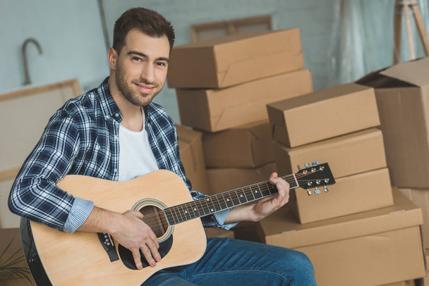 portrait of smiling man with acoustic guitar at new home with cardboard boxes, relocation concept - Photo, Image