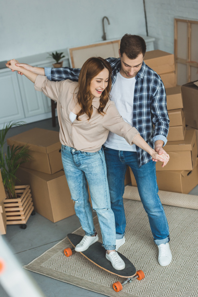 young couple having fun with skateboard at new apartment with cardboard boxes, relocation concept - Photo, Image