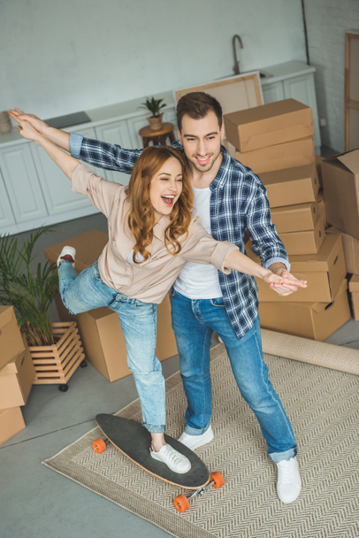 young couple having fun with skateboard at new apartment with cardboard boxes, relocation concept - Photo, Image