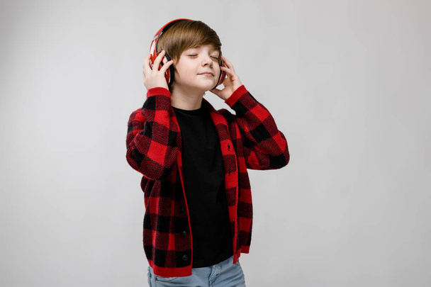 Kedves preteen kaukázusi fiú alkalmi ruhában piros fülhallgatóval zenét hallgatni, és bemutatja a különböző kifejezések fehér falon a stúdió. - Fotó, kép
