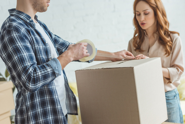 partial view of couple packing cardboard box with sticky tape together, relocation concept - Photo, Image