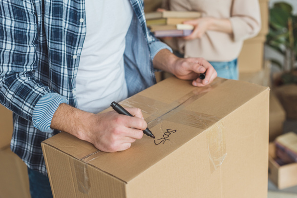 cropped shot of man signing cardboard box with wife with books in hands near by, moving home concept - Photo, Image