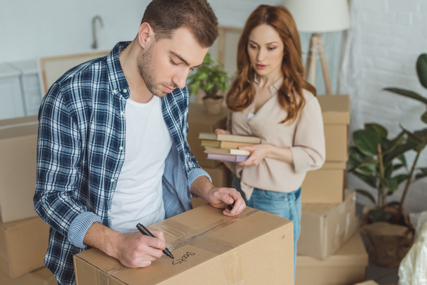 man signing cardboard box with wife with books in hands near by, moving home concept - Photo, Image