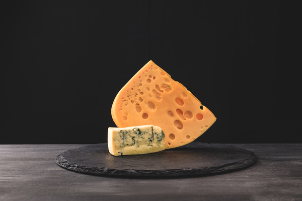 Closeup view of emmental and blue cheese on board on black  - Zdjęcie, obraz