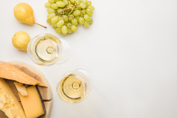 Top view of different types of cheese on wooden board, wine glasses and fruits on white - Photo, Image