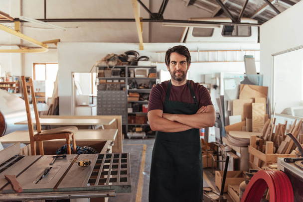 Portrait of skilled young woodworker with beard, standing confidently with his arms crossed by bench saw in his workshop full of carpentry equipment - Foto, Imagem