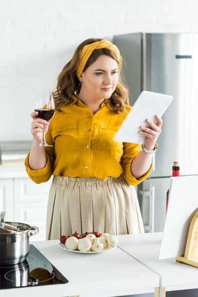 beautiful woman holding glass of red wine and using tablet in kitchen - Фото, зображення