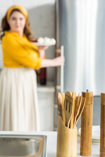 woman taking plate from fridge with wooden spatulas on foreground in kitchen - Photo, Image