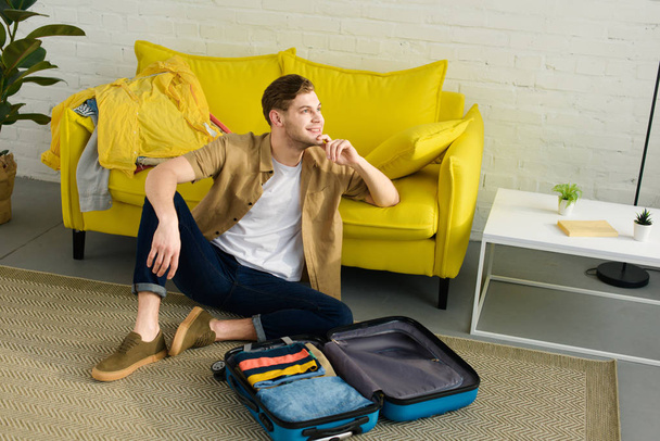 красивий мрійливий чоловік сидить на підлозі біля дорожньої сумки
 - Фото, зображення