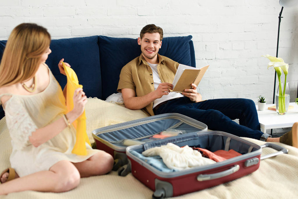 pregnant woman packing clothes for vacation while husband reading book on bed - Photo, Image
