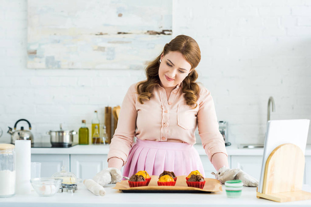 smiling beautiful woman looking at tray with muffins at kitchen - Valokuva, kuva