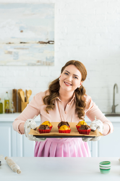 mosolygó gyönyörű nő betöltő tálca muffint és látszó-on fényképezőgép-konyha - Fotó, kép