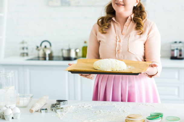 imagen recortada de mujer sosteniendo bandeja con masa para hornear pan en la cocina
 - Foto, Imagen
