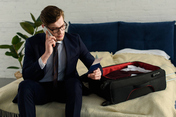 handsome businessman holding air ticket with passport and talking on smartphone while sitting on bad with suitcase - Photo, Image