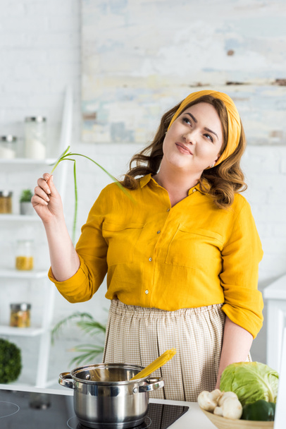 pensive beautiful woman holding green onion in kitchen - Photo, Image
