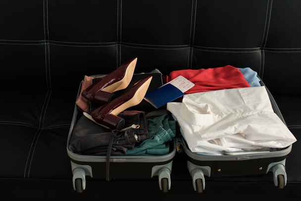 opened travel bag with passport, clothes and heels on sofa - Foto, imagen