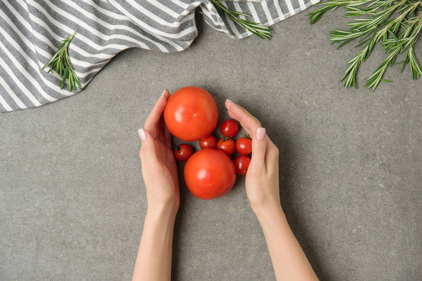 tiro recortado de manos femeninas sosteniendo tomates maduros en la superficie de hormigón gris con lino y romero
 - Foto, imagen
