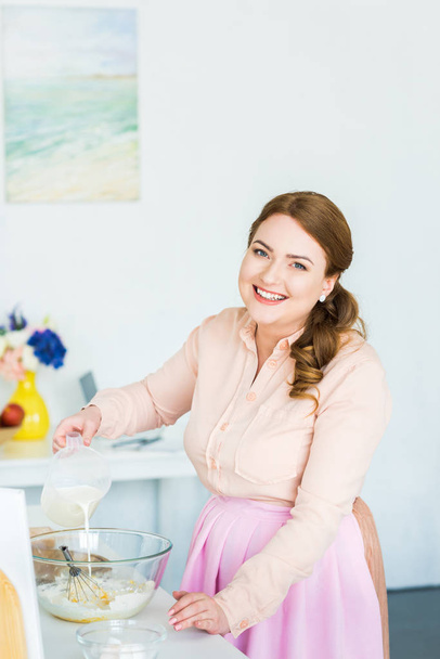 sonriente hermosa mujer verter leche en un tazón con harina en la cocina
 - Foto, Imagen