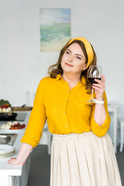 cheerful beautiful woman holding glass of wine and looking away in kitchen - 写真・画像