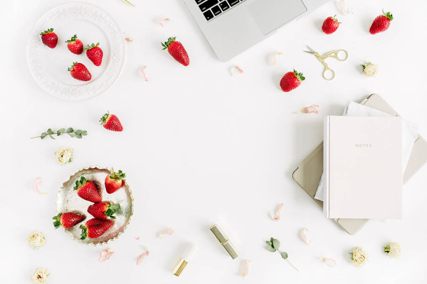 Female modern home office desk with laptop, notebook, lipstick, fresh raw strawberries and rose flower buds on white background. Flat lay, top view. Frame with copy space for text. - Photo, Image