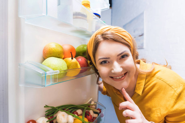 Lächelnde schöne Frau blickt in die Kamera in der Nähe des offenen Kühlschranks in der Küche - Foto, Bild