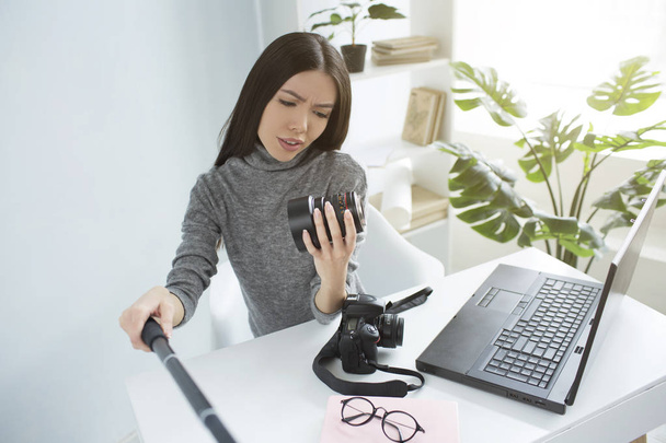 Professional blogger is sitting at the table and recording a video where she is looking to the cameras lens and studying it. She looks serious and cocncentrated. - 写真・画像