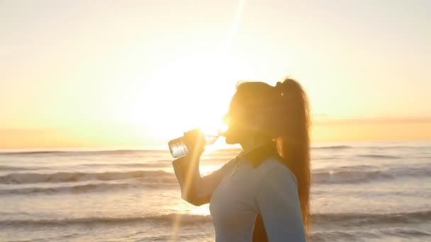 Жінка пити з пляшки води на заході сонця біля моря - Кадри, відео