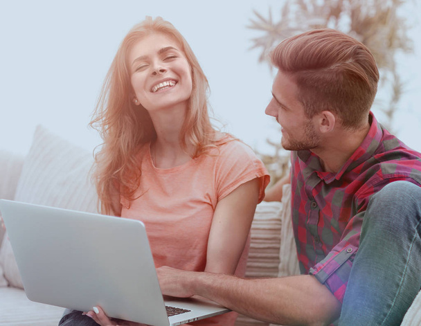 young couple is using a laptop and smiling while sitting on sofa at home - Φωτογραφία, εικόνα