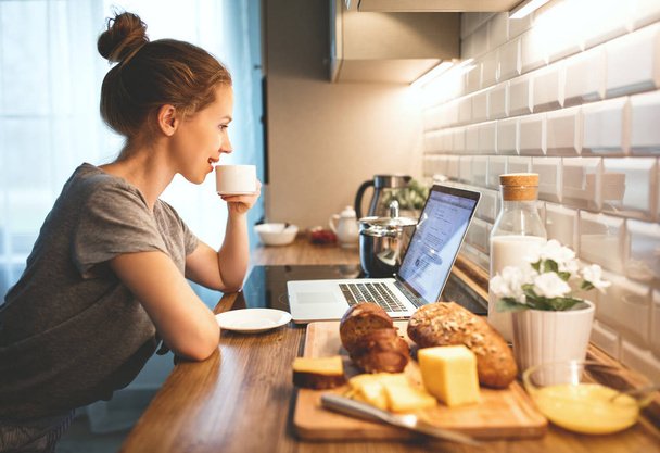 Frau im Schlafanzug frühstückt in Küche am Computer - Foto, Bild