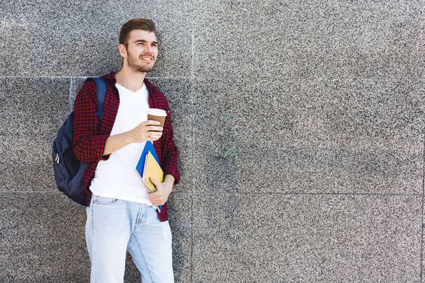 Красивый студент держит чашку кофе на сером фоне на открытом воздухе
 - Фото, изображение