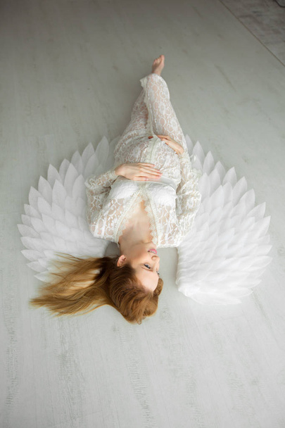 beautiful young pregnant girl with red hair in a lacy white dress lying on a light floor - Фото, зображення