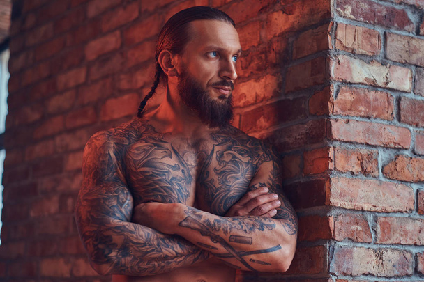Ένας όμορφος tattoed shirtless αρσενικό με ένα μοντέρνο κούρεμα και γενειάδα, στέκεται με σταυρωμένα χέρια, κλίνει ενάντια σε έναν τοίχο από τούβλα σε ένα δωμάτιο με ένα εσωτερικό πατάρι. - Φωτογραφία, εικόνα