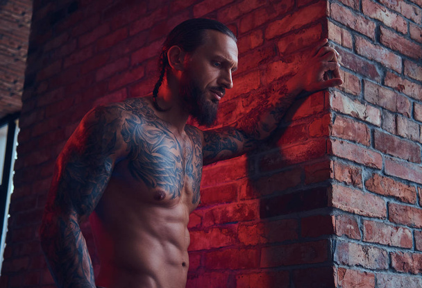 Een knappe tattoed shirtless man met een stijlvol kapsel en baard, staande tegen een bakstenen muur in een kamer met een loft interieur. - Foto, afbeelding