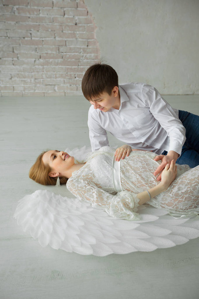 beautiful couple of a man and a pregnant woman in a white lace dress with wings lying on a light floor - Zdjęcie, obraz