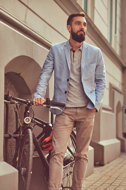 おしゃれなひげを生やした男性の身に着けているスタイリッシュな服、アンティークのヨーロッパの通りの自転車スタンド. - 写真・画像