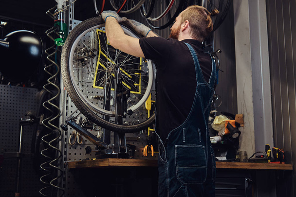 Красивый рыжий мужчина в джинсовом комбинезоне, работает с велосипедным колесом в ремонтной мастерской. Рабочий снимает велосипедную шину в мастерской
. - Фото, изображение